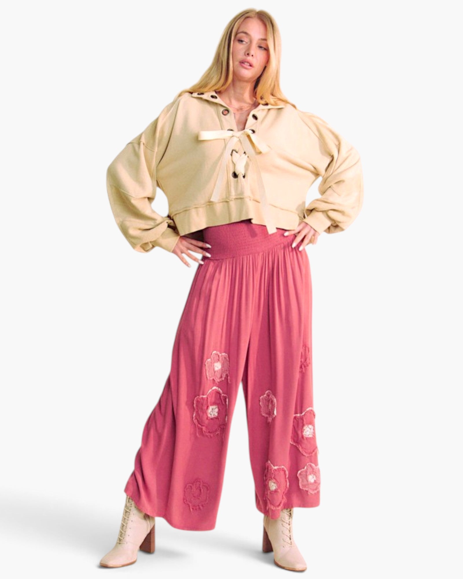 A model wearing a beige cropped tie-front jacket and pink wide-leg pants with floral embroidery, posed against a light gray studio background.
