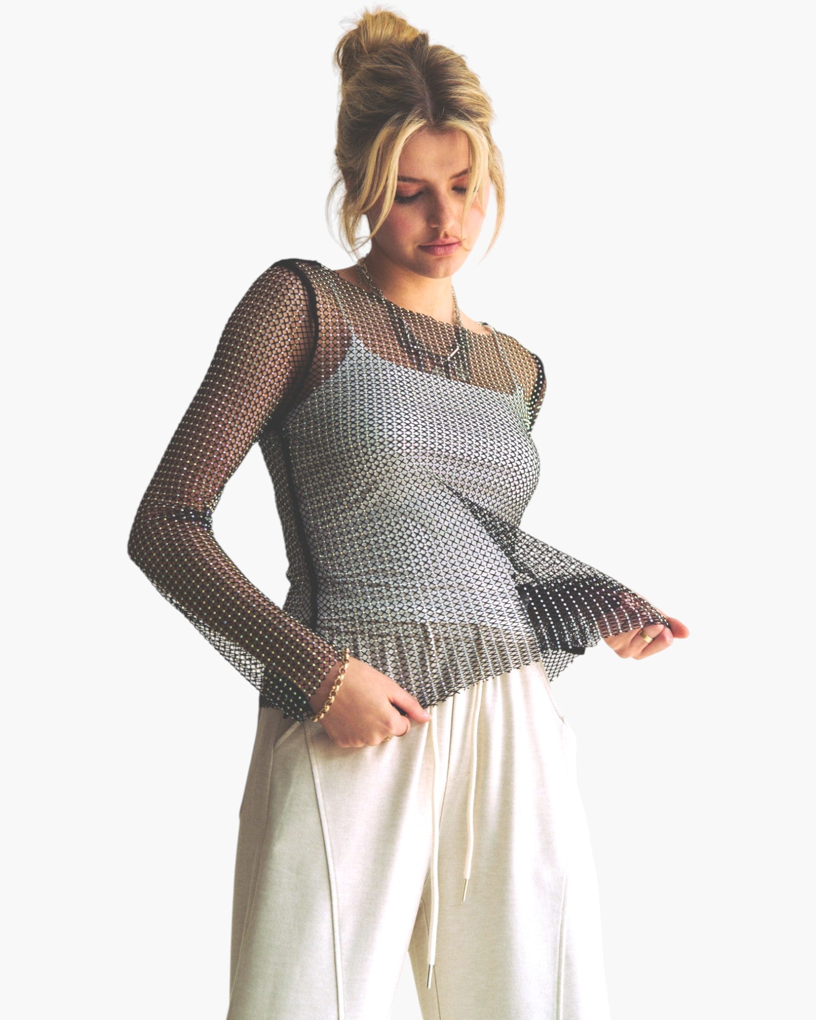 A woman modeling a sheer black mesh long-sleeve top over a silver patterned camisole, paired with off-white drawstring pants, against a plain white background.