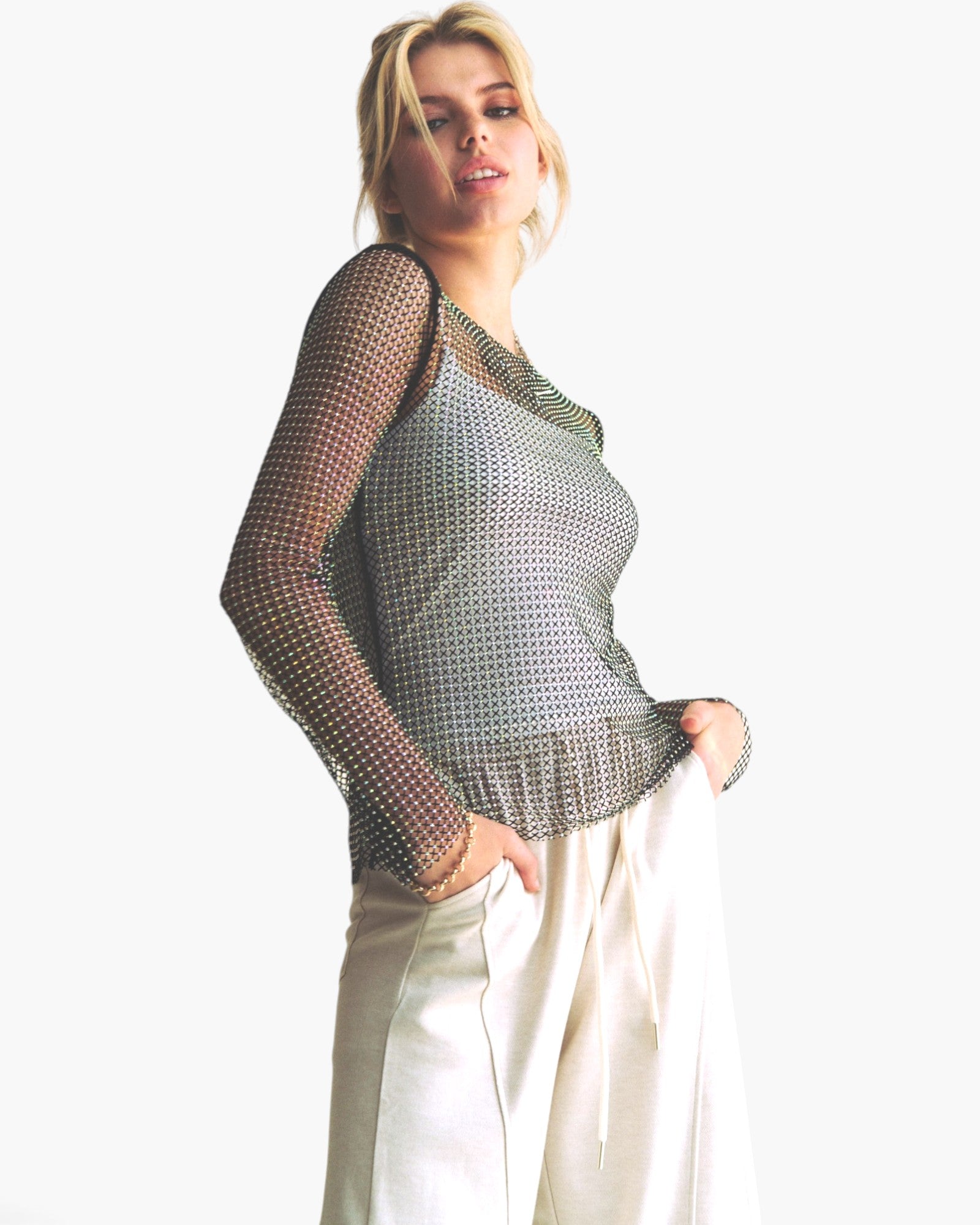 A woman modeling a silver mesh long-sleeve top with a scoop neckline, paired with cream-colored pants, against a plain white background.