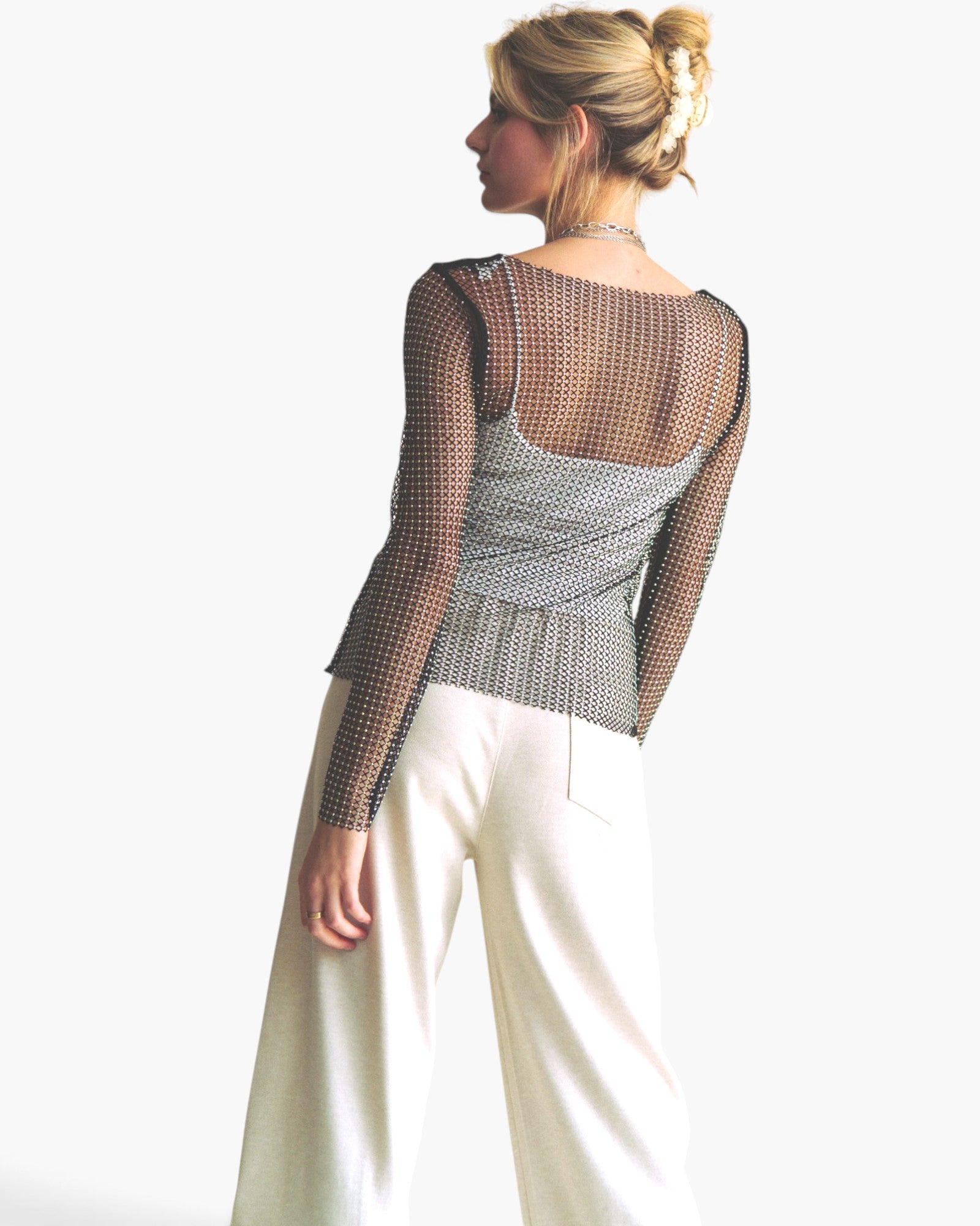 A woman shown from the back wearing a sheer black mesh long-sleeve top over a gray patterned camisole, paired with white wide-leg pants, against a plain white background.