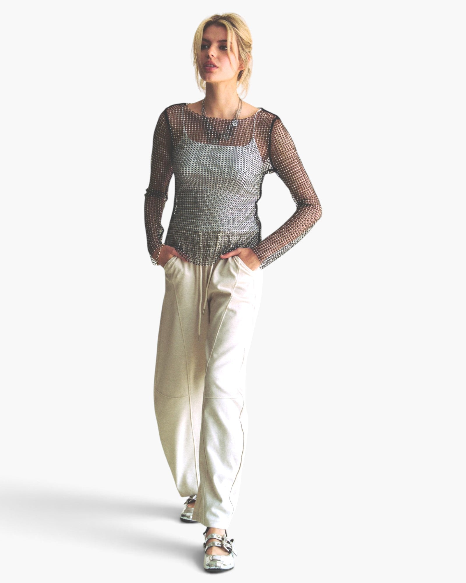 A woman modeling a sheer black mesh long-sleeve top with a silver and black checkered bodice, paired with cream-colored wide-leg pants and metallic sandals, against a plain white background.
