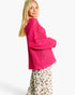 A blonde woman wearing a bright pink open-knit cropped sweater and leopard-print pants, photographed against a clean white studio background.
