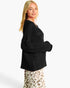 A woman modeling a black textured knit sweater with a wide collar and long sleeves, paired with a leopard-print skirt, against a plain white studio background.