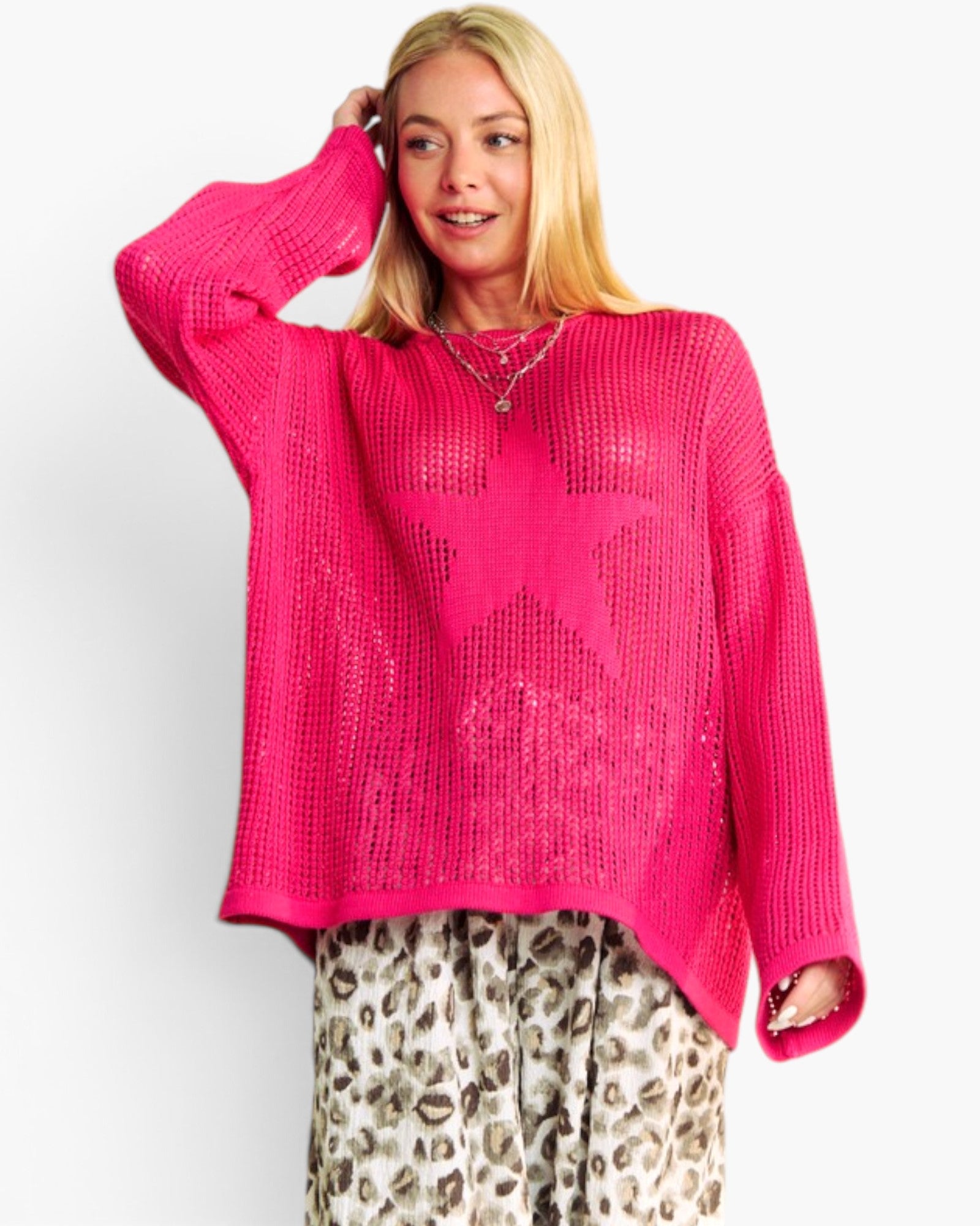 A woman modeling a bright pink knit sweater with a large star motif on the front, paired with leopard-print pants, against a plain light gray background.