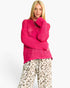 A blonde model wearing a bright pink chunky knit sweater and beige leopard-print pants, standing against a plain white studio background.
