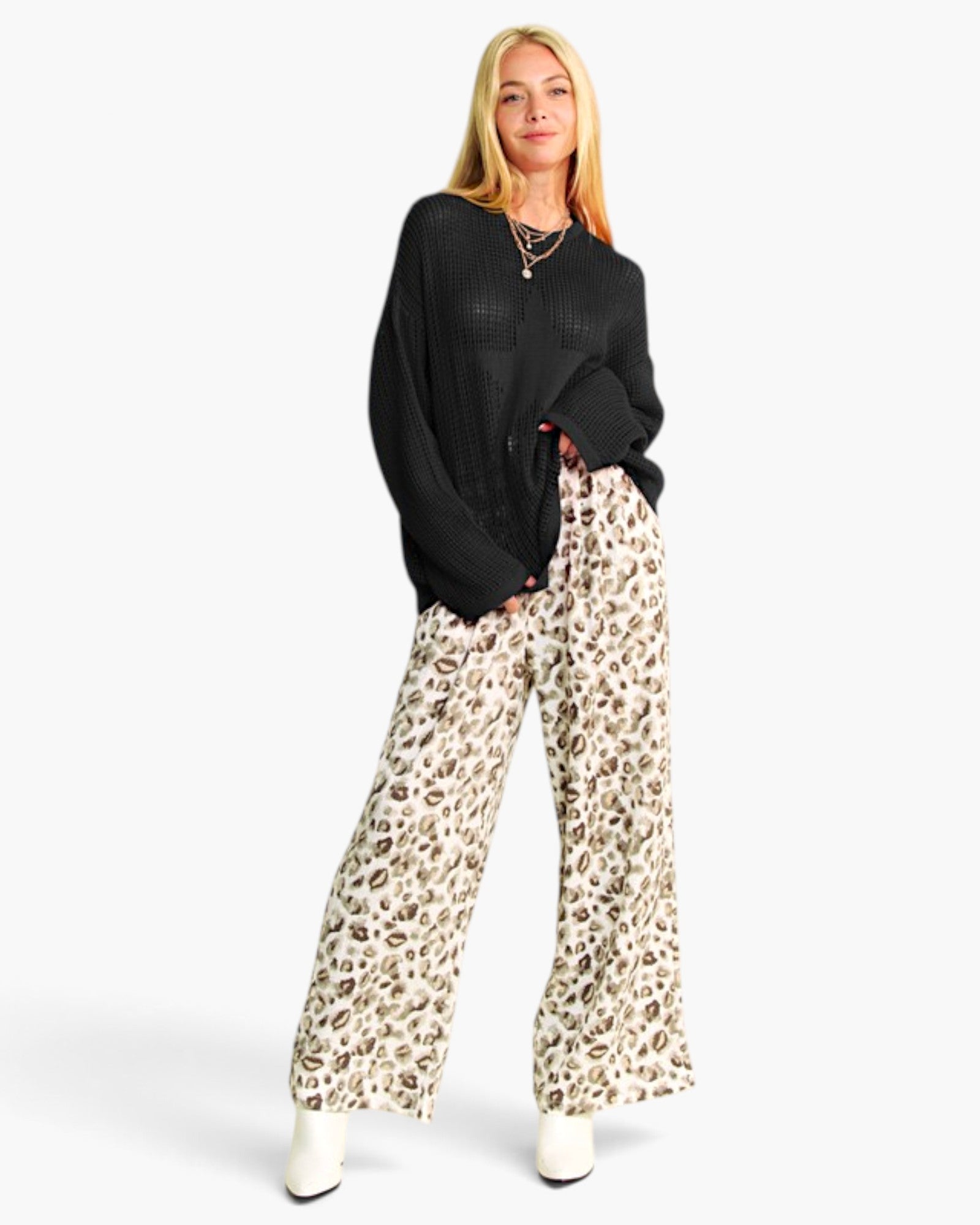 A woman modeling a black knit long-sleeve top and wide-leg beige pants with a brown leopard print, paired with white boots, against a light gray studio background.