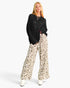 A woman modeling a black knit long-sleeve top and wide-leg beige pants with a brown leopard print, paired with white boots, against a light gray studio background.