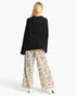 A woman shown from the back wearing a black textured knit sweater and wide-leg beige pants with a brown and black leopard print, paired with white high heels, against a light gray studio background.