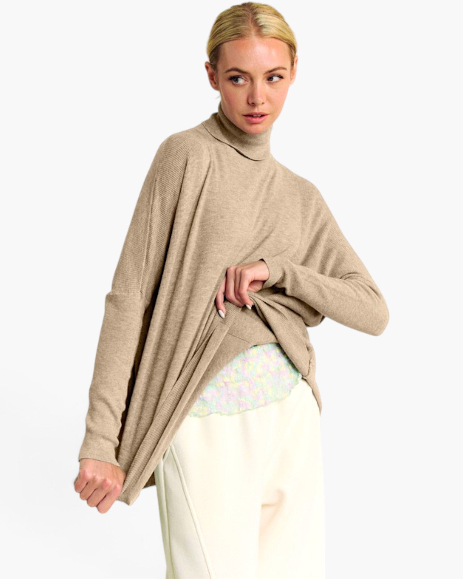 A woman modeling a beige, draped turtleneck sweater over a pastel patterned top and cream pants, set against a plain light gray studio background.