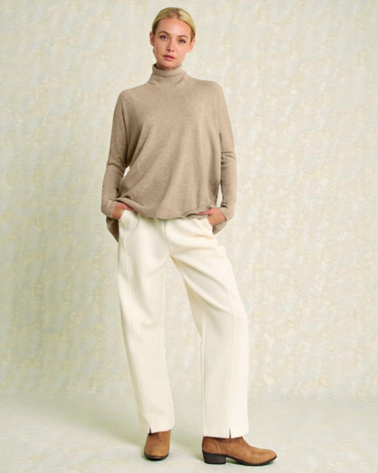 Person wearing a beige turtleneck sweater and white pants on a light background