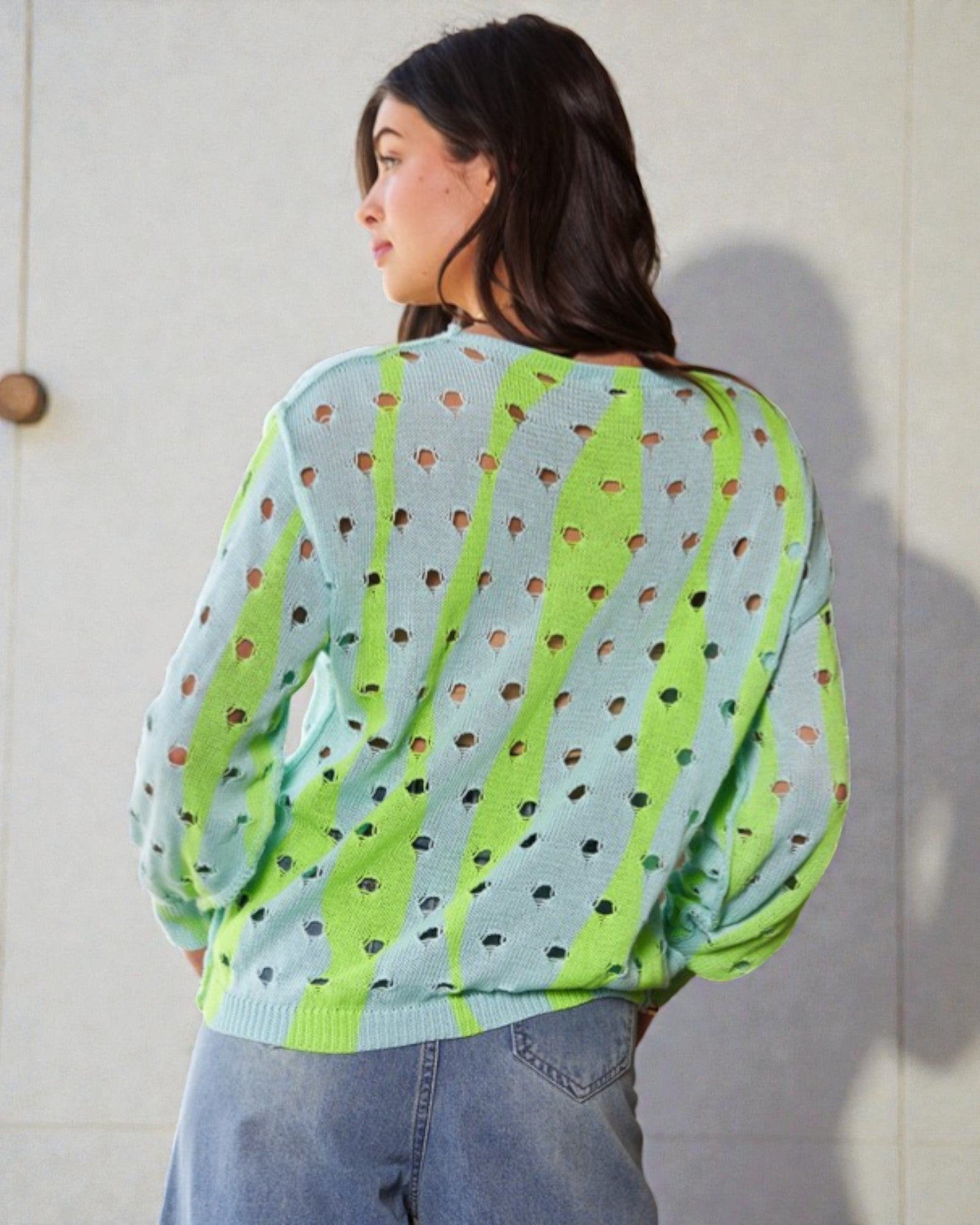 Person wearing a light blue sweater with green stripes and black dots, standing against a white wall.