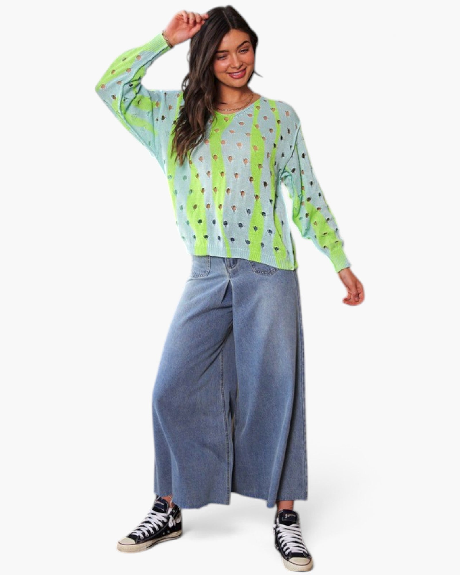 A woman modeling a long-sleeve knit top with a lime green and light blue gradient and small colorful geometric patterns, paired with wide-leg denim pants and black sneakers against a plain white background.