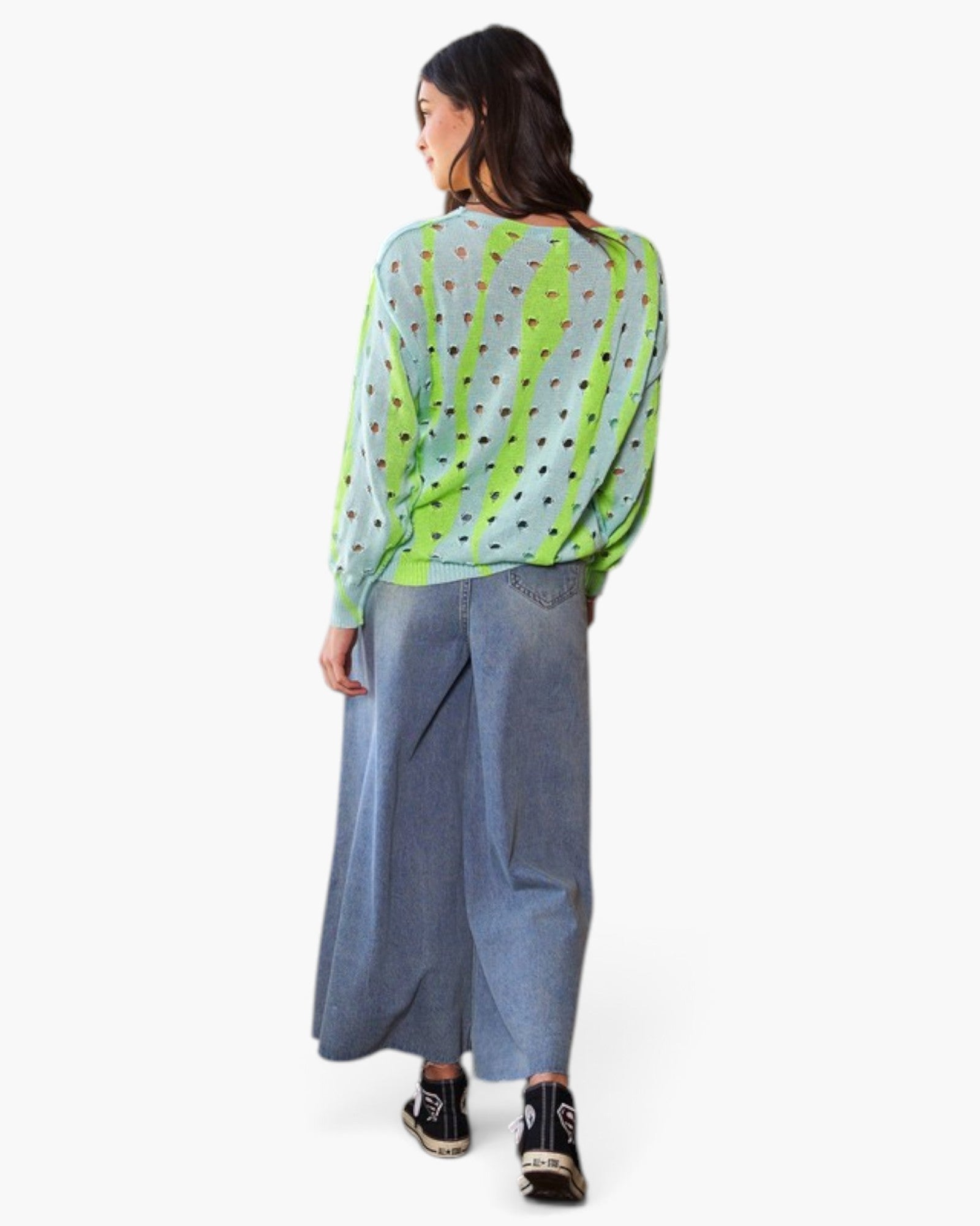 A woman is shown from behind wearing a sheer light blue-green long-sleeve top with vertical neon green stripes and black polka dots, paired with wide-leg blue jeans and black sneakers, against a plain white background.