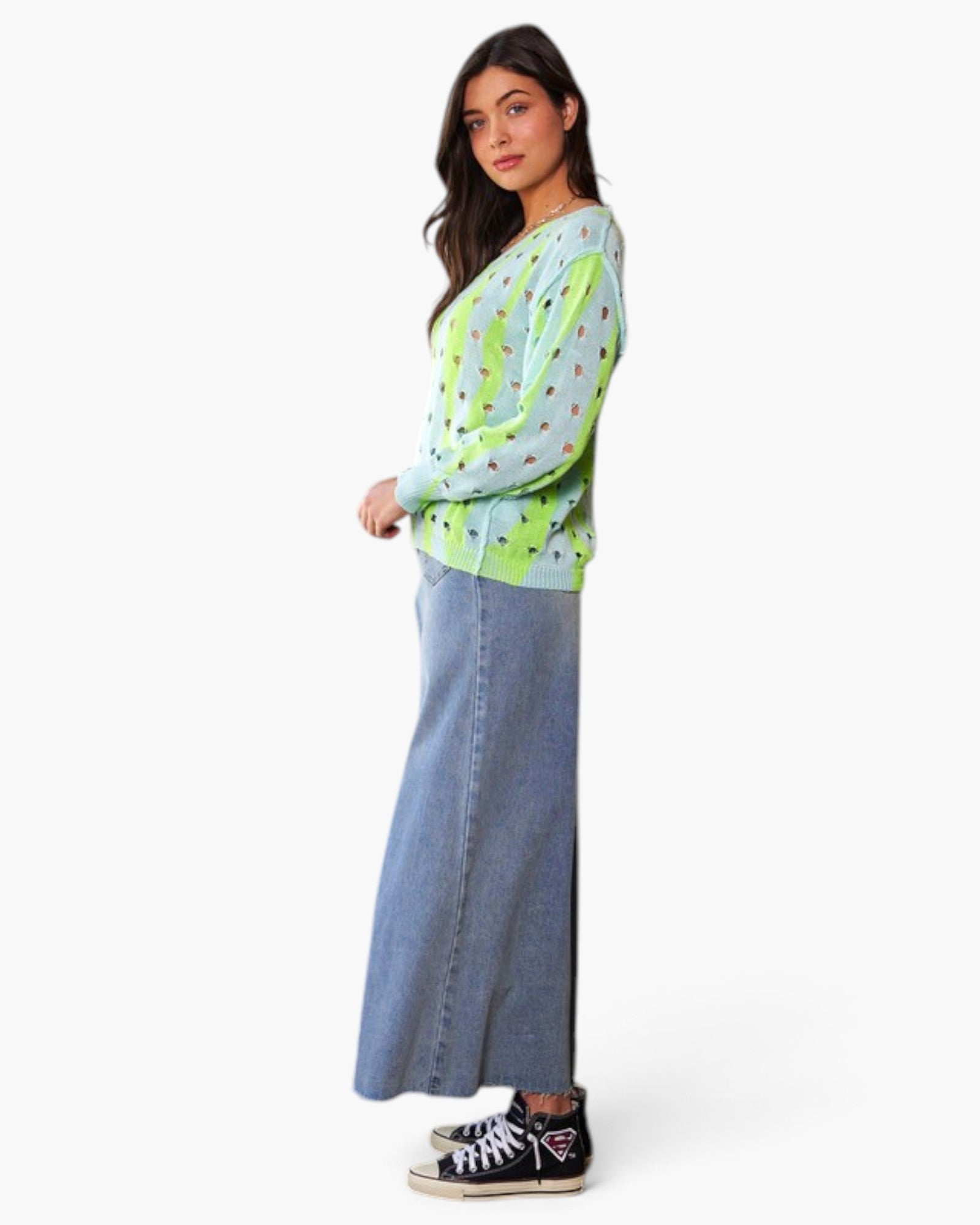 A woman modeling a light green knit cardigan with a colorful polka-dot pattern, paired with a wide-leg blue denim skirt and black high-top sneakers, against a plain white studio background.
