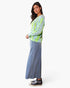 A woman modeling a light green knit cardigan with a colorful polka-dot pattern, paired with a wide-leg blue denim skirt and black high-top sneakers, against a plain white studio background.