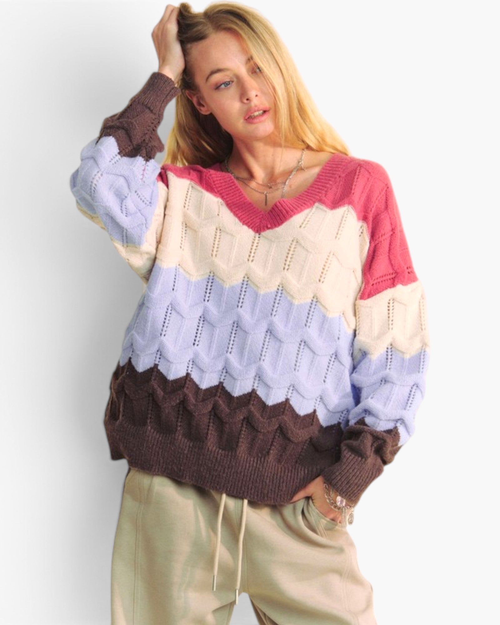 A woman wearing a chunky knit sweater with horizontal color-blocked stripes in pink, cream, light blue, and dark brown, paired with beige drawstring sweatpants against a light gray studio background.