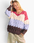 A woman wearing a chunky knit sweater with horizontal color-blocked stripes in pink, cream, light blue, and dark brown, paired with beige drawstring sweatpants against a light gray studio background.