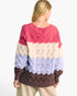 A woman shown from the back wearing a chunky knit sweater with horizontal color-blocked stripes in pink, cream, light blue, and dark brown, paired with beige pants against a plain white background.