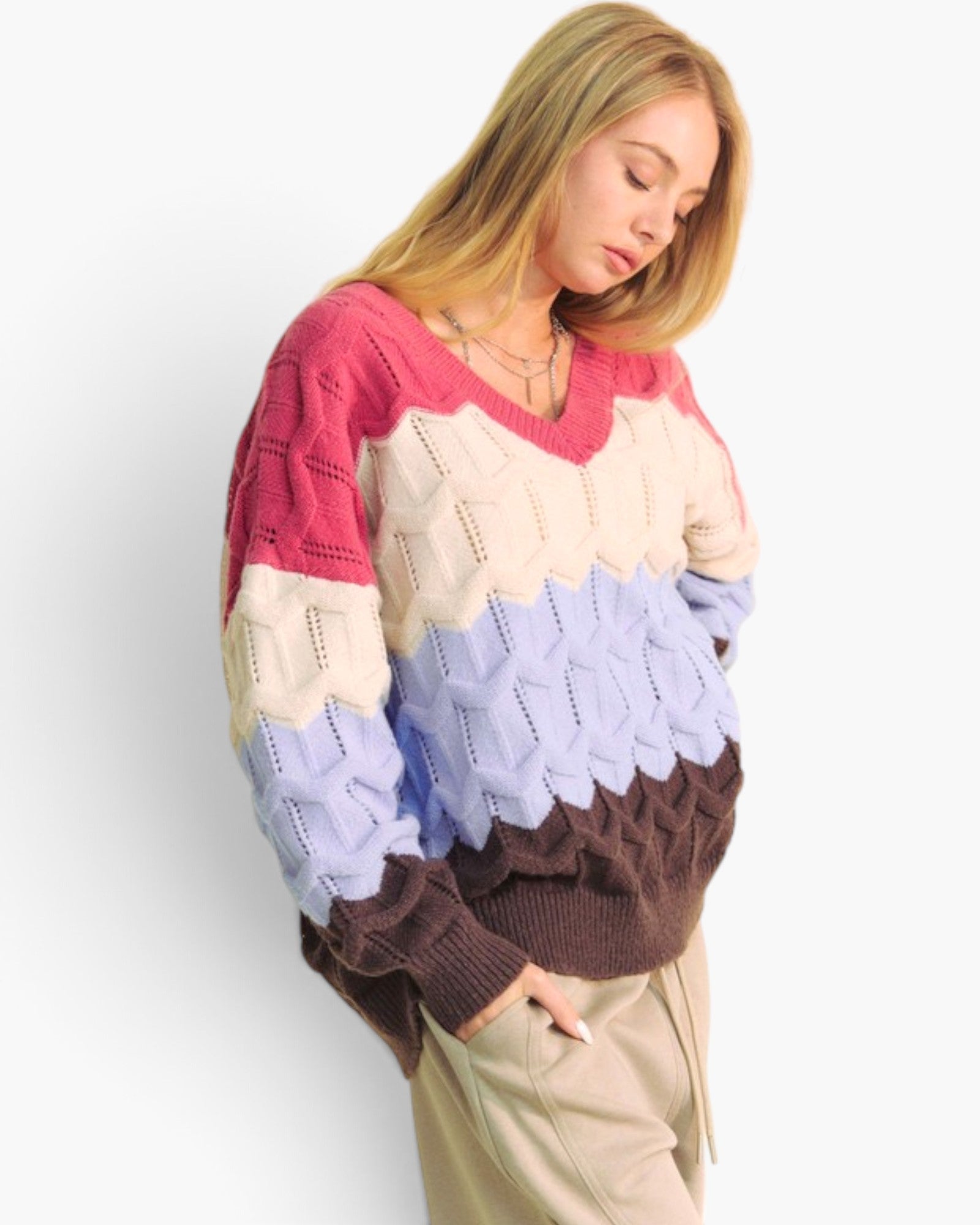 A woman wearing a chunky color-block knit sweater with pink, cream, light blue, and brown sections, paired with beige pants, against a plain white background.
