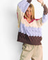 A woman modeling a chunky knit sweater with horizontal color-blocked stripes in lavender, cream, and dark brown, paired with beige drawstring pants against a plain white studio background.