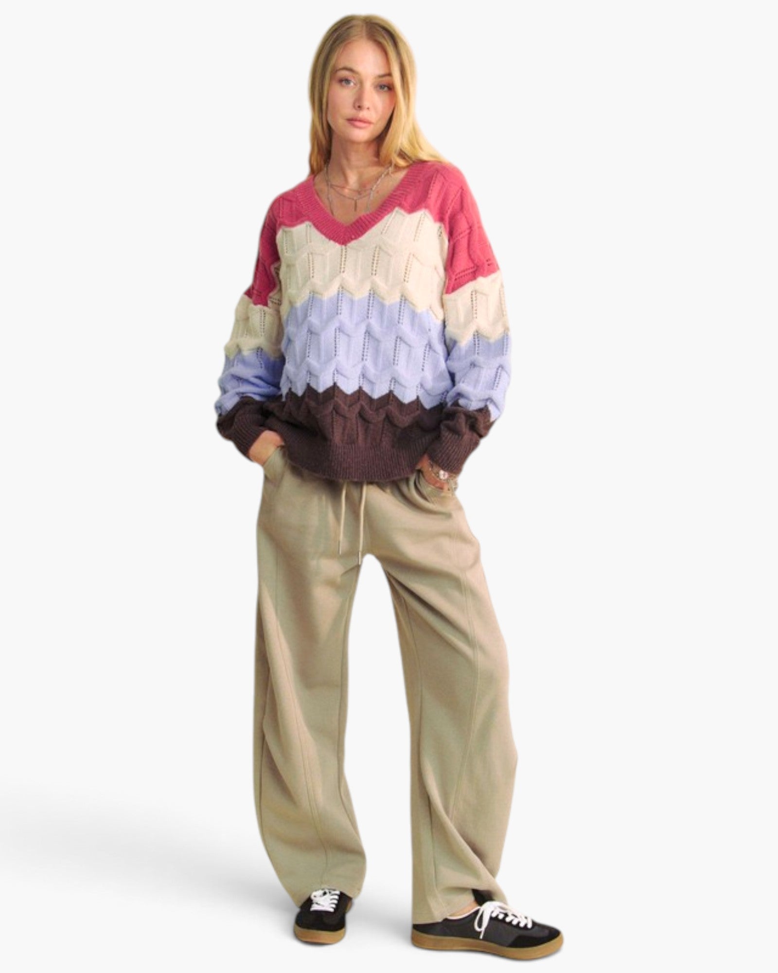 A woman modeling a chunky knit color-block sweater with pink, cream, lavender, and dark brown horizontal stripes, paired with beige drawstring pants and black sneakers, against a plain white studio background.