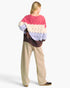 A woman shown from the back wearing a chunky cable-knit sweater with horizontal color-blocked stripes in pink, cream, light blue, and dark brown, paired with beige wide-leg pants and black slip-on shoes, against a plain white studio background.