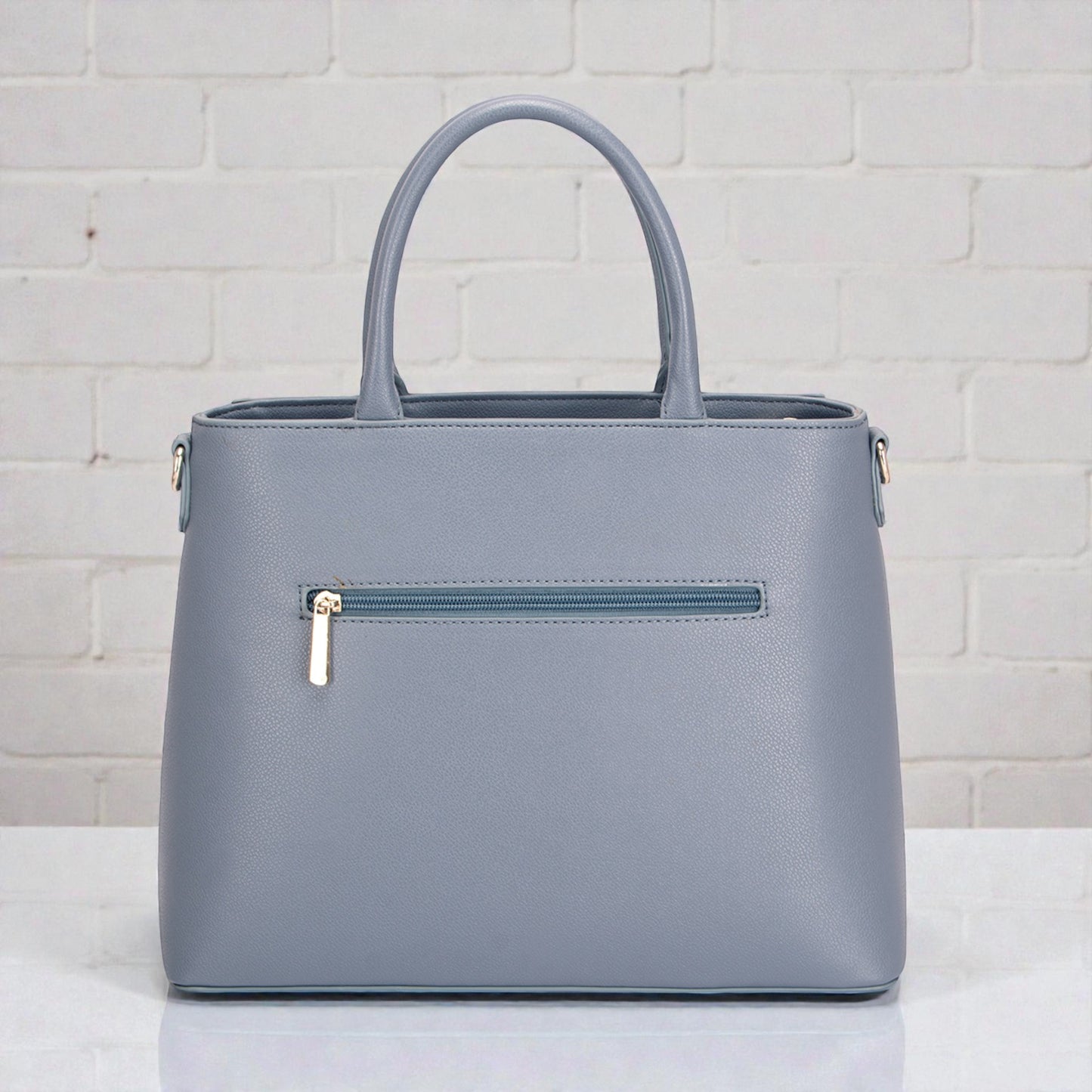 Gray handbag with a gold zipper on a white surface and light gray brick wall background