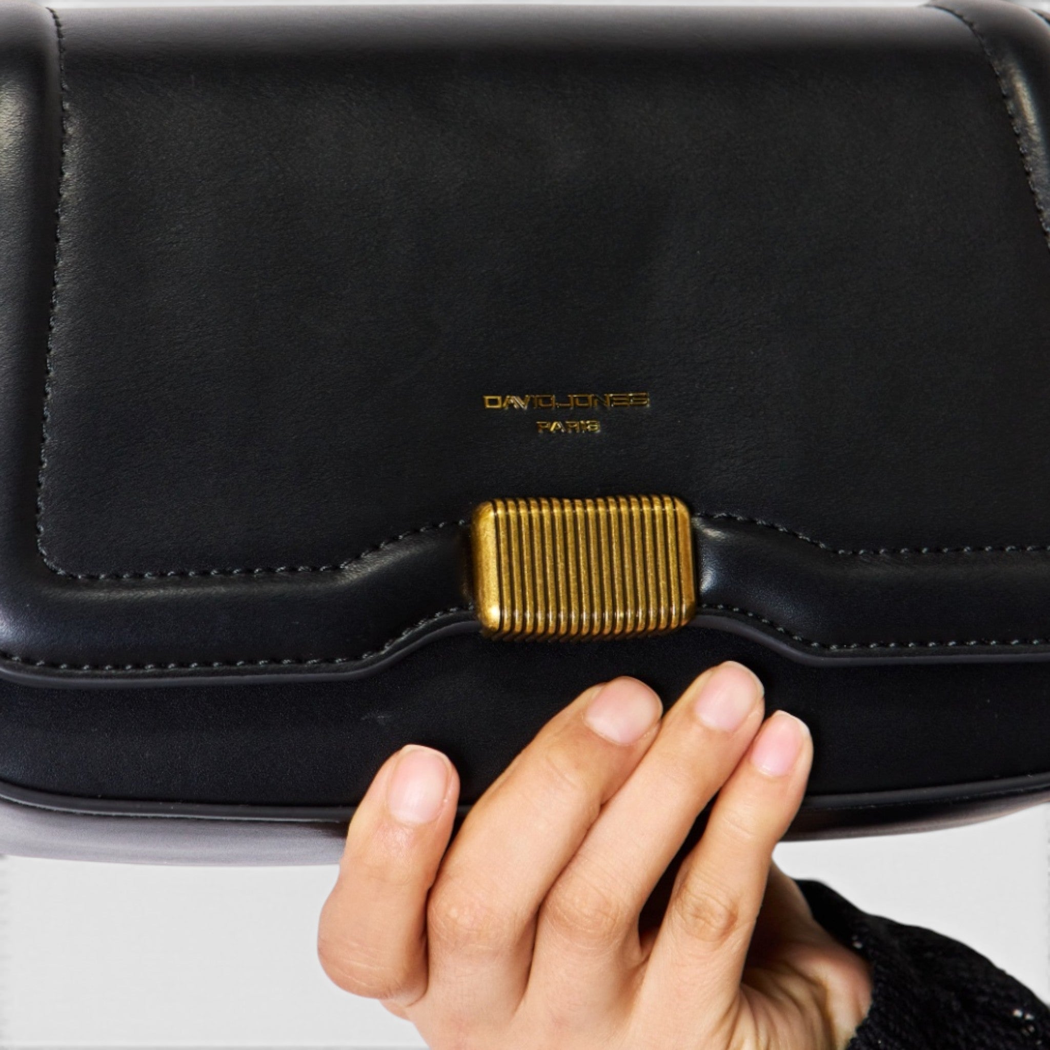 Black clutch bag with gold clasp held by a hand on a light gray background
