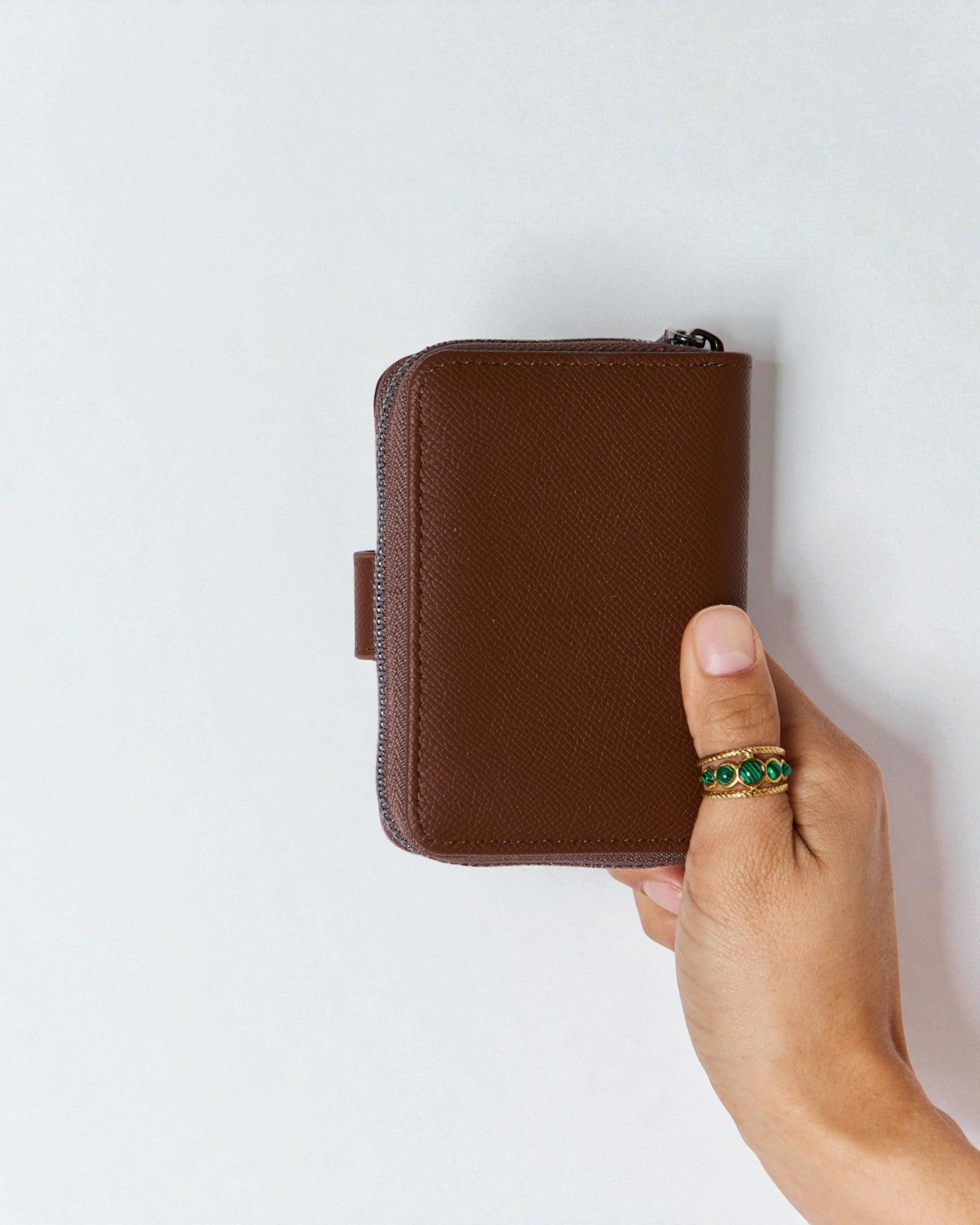 Brown wallet held by a hand with a green ring on a white background