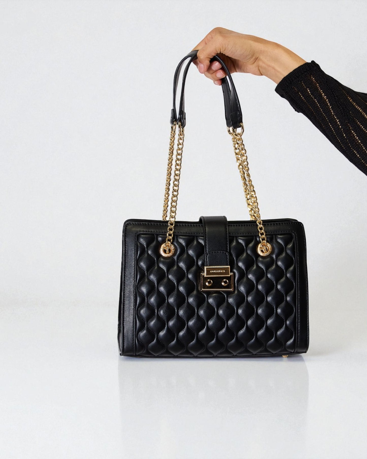 Black quilted handbag with gold chain handle held by a hand on a white background