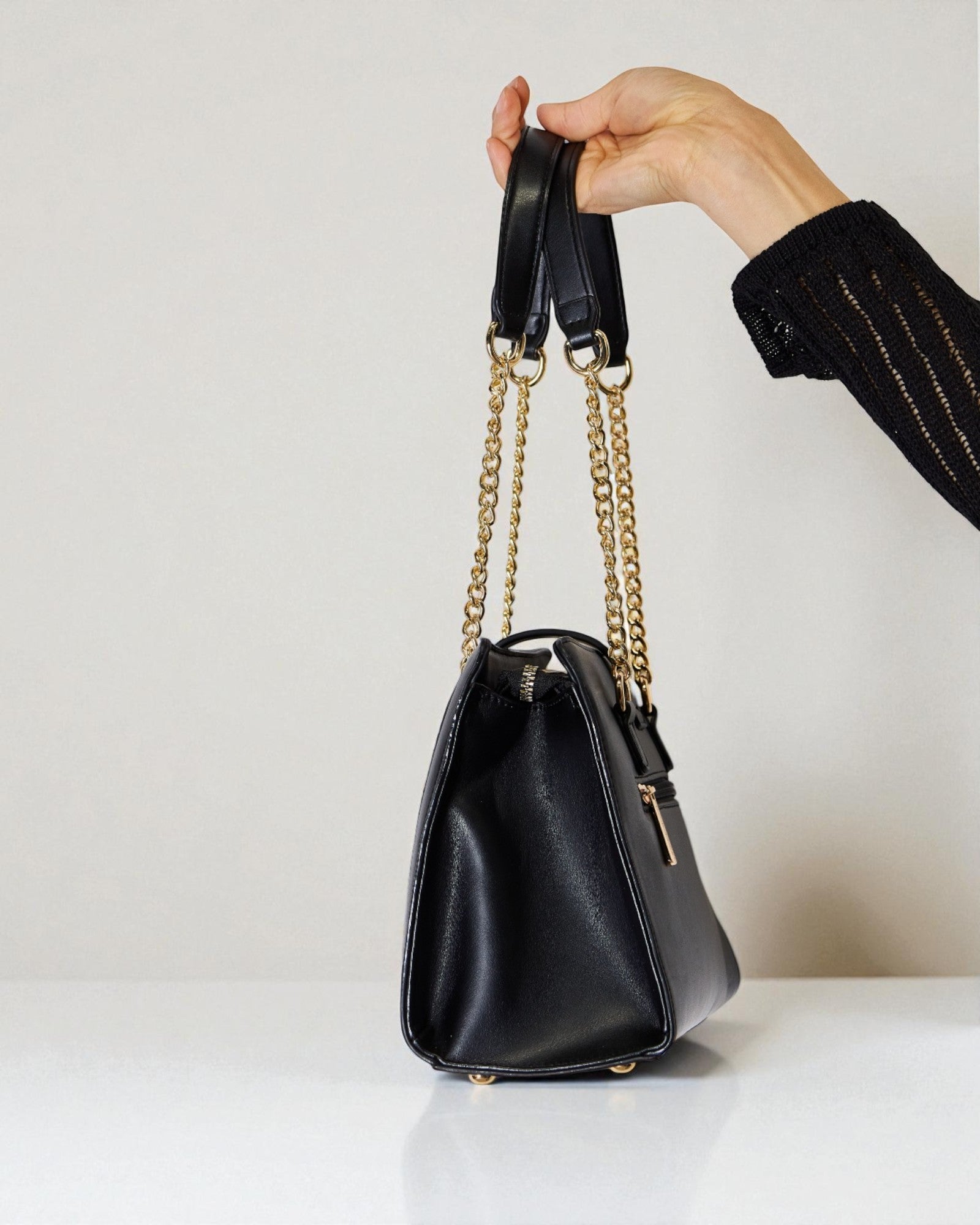 Black handbag with gold chain strap held by a hand against a neutral background