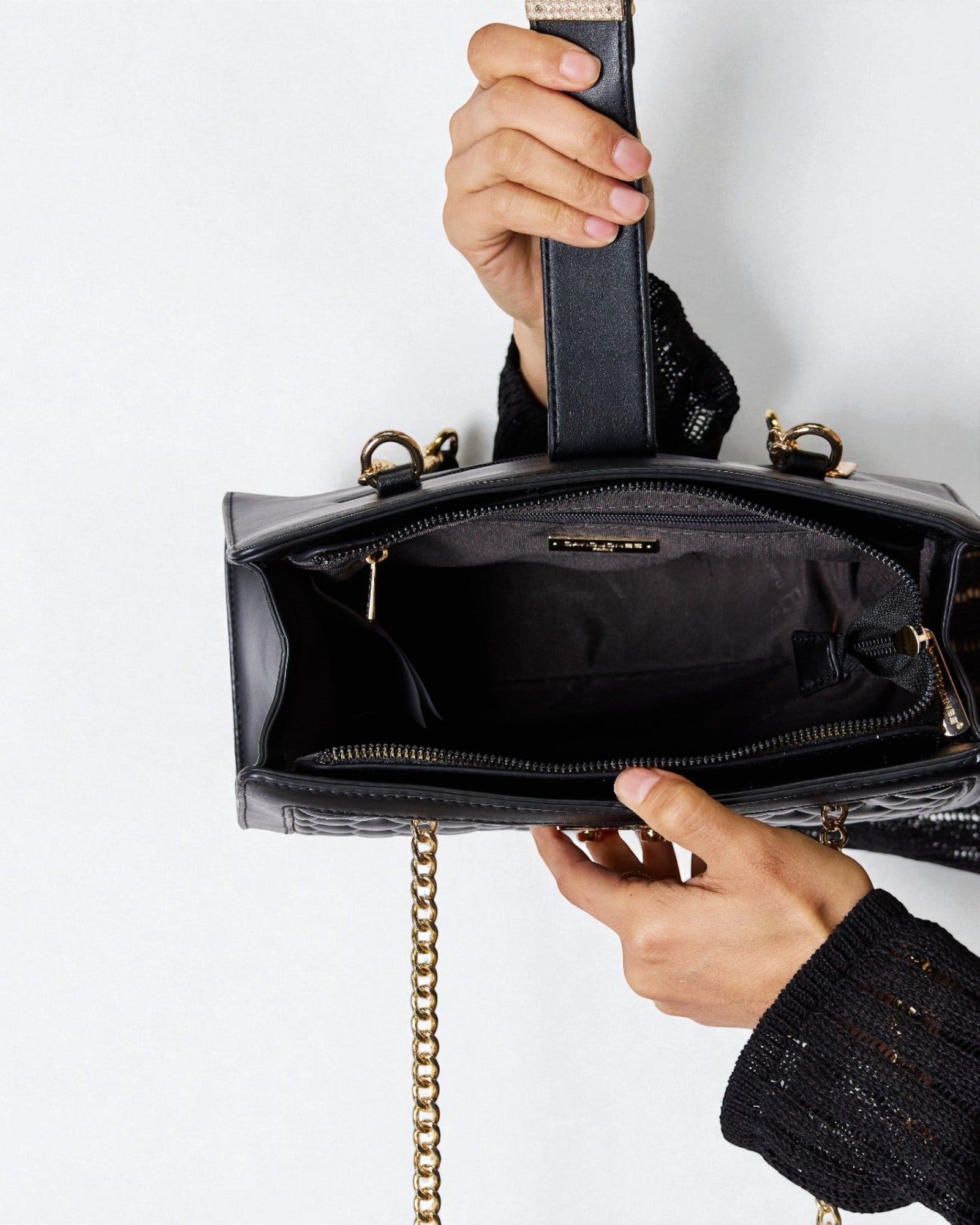 Black handbag with gold chain strap held by a person against a white background
