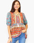 A woman wearing a colorful bohemian-style short-sleeve top with a tassel tie neckline, paired with blue jeans, against a plain white background.