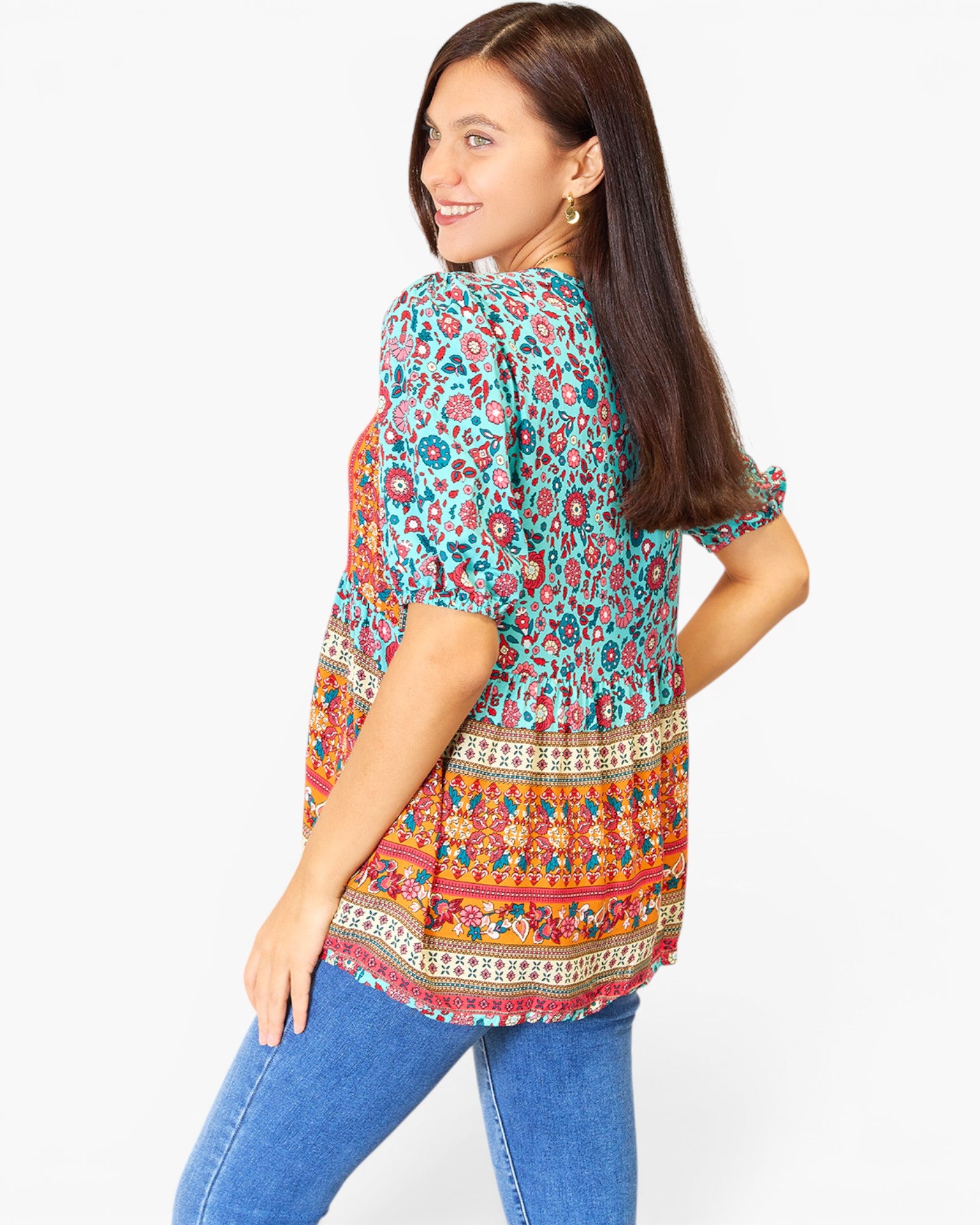 A woman wearing a colorful short-sleeve tunic top with a floral and geometric print, paired with blue jeans, photographed against a plain white background.