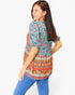 A woman wearing a colorful short-sleeve tunic top with a floral and geometric print, paired with blue jeans, photographed against a plain white background.