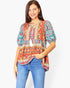 A woman wearing a colorful bohemian-style short-sleeve top with blue jeans, standing against a plain white studio background.