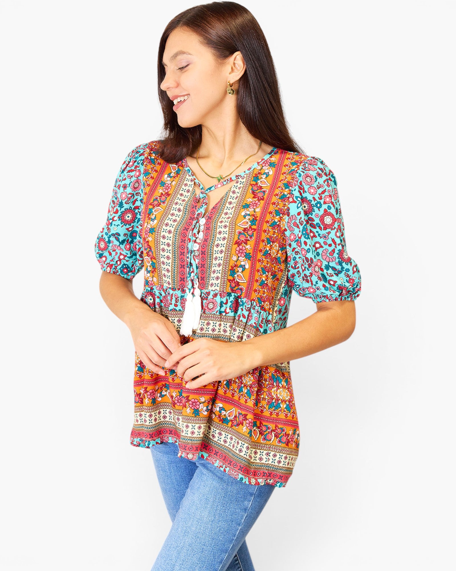 A woman wearing a colorful bohemian-style short-sleeve blouse with blue jeans, standing against a plain white studio background.