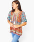 A woman wearing a colorful bohemian-style short-sleeve blouse with blue jeans, standing against a plain white studio background.