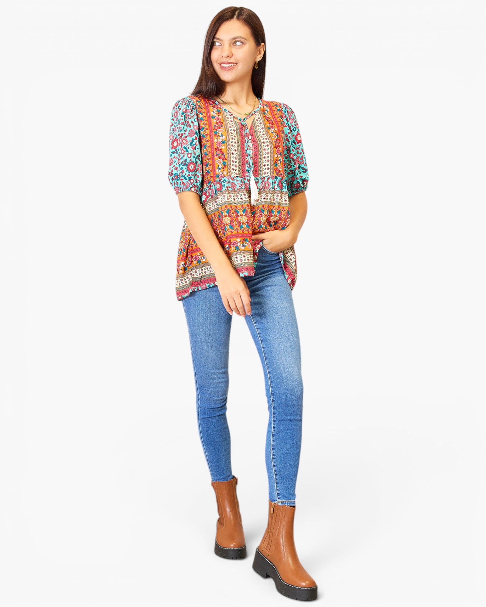 A woman modeling a short-sleeve bohemian-style top with a colorful geometric and floral print, paired with blue skinny jeans and brown ankle boots, against a plain white background.