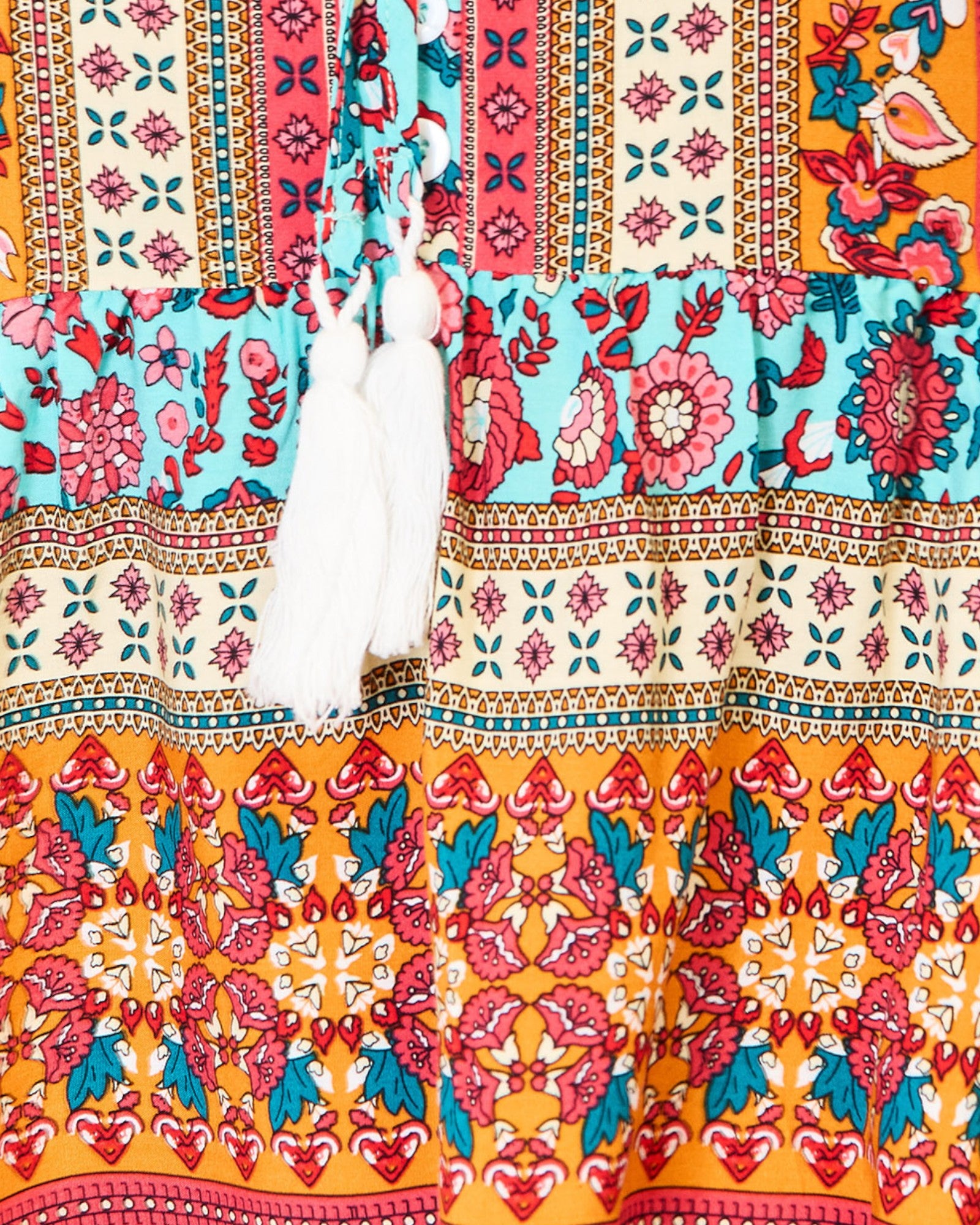 A close-up of a colorful patterned garment, likely a bohemian-style blouse or dress, featuring tiered sections with white tassels at the neckline. The fabric displays multiple vibrant patterns including floral prints on turquoise, pink, orange, and cream, separated by geometric borders.