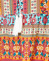 A close-up of a colorful patterned garment, likely a bohemian-style blouse or dress, featuring tiered sections with white tassels at the neckline. The fabric displays multiple vibrant patterns including floral prints on turquoise, pink, orange, and cream, separated by geometric borders.