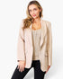 A woman modeling a light beige blazer over a beige cable-knit top and black pants, photographed against a clean white studio background.