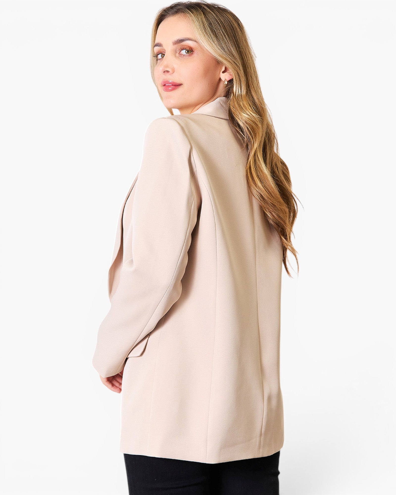 A woman with long blonde hair is shown from the back wearing a light beige blazer over black pants, photographed against a clean white studio background.