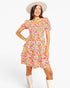 A woman modeling a short-sleeve, ruffled mini dress with a vibrant multicolor floral print on a white background, styled with a cream wide-brim hat and cream boots against a light gray studio background.