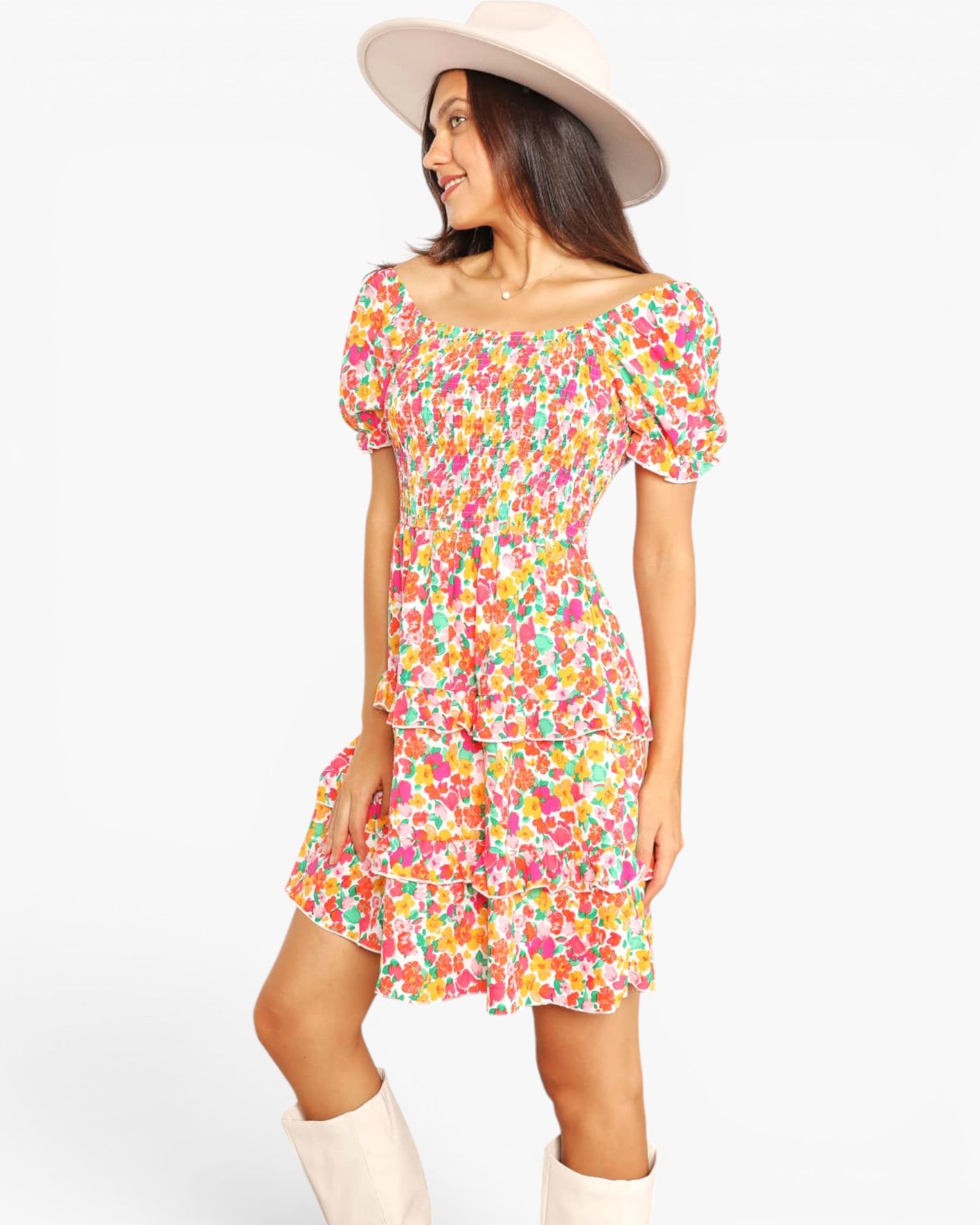 A woman modeling a short, off-the-shoulder dress with a colorful floral print, paired with a wide-brimmed cream hat and cream boots, against a plain white background.