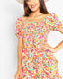 A woman wearing a colorful off-the-shoulder dress with a vibrant floral print in pink, orange, yellow, green, and blue on a white base, paired with a light-colored wide-brim hat against a plain light gray background.