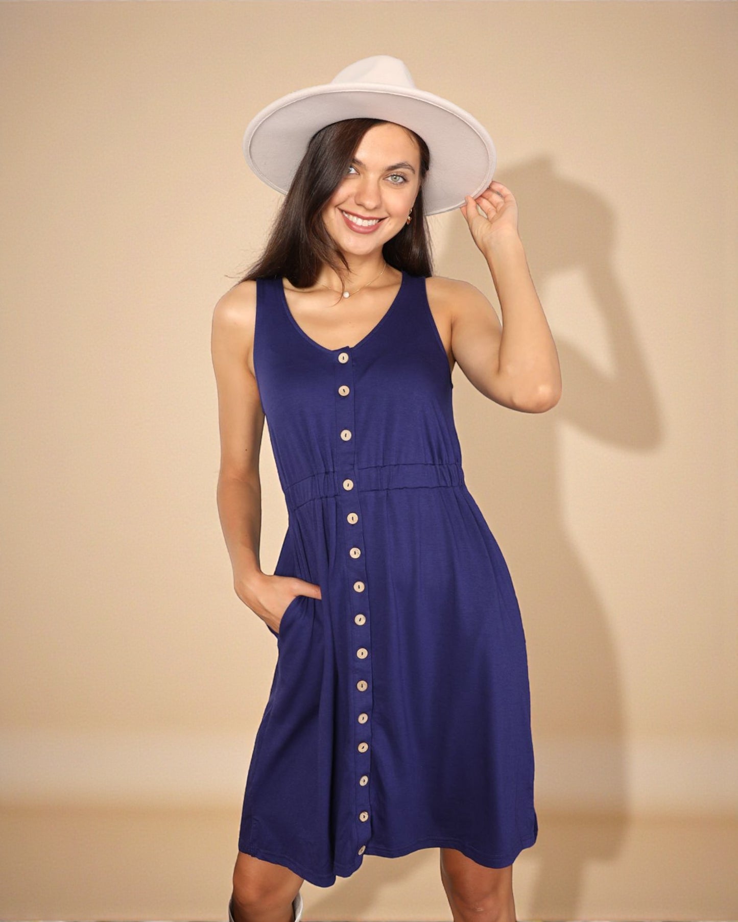 Woman wearing a blue dress with buttons and a white hat against a beige background