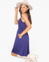 A woman modeling a sleeveless, scoop-neck blue dress with side pockets, paired with a beige wide-brim hat and beige knee-high boots, set against a plain white studio background.