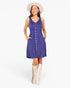 A woman modeling a sleeveless blue-purple A-line dress with a button-down front, side pockets, and a gathered waist, styled with a cream wide-brim hat and cream knee-high boots against a light gray studio background.