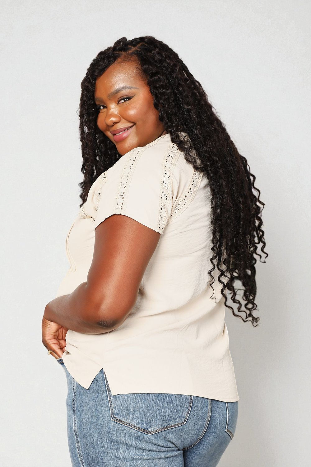 Woman with long, curly hair wearing a beige top and blue jeans against a white background